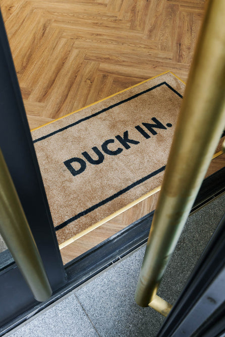 Duck & Dry Mayfair salon doorway with a "Duck In." mat on the herringbone wood floor