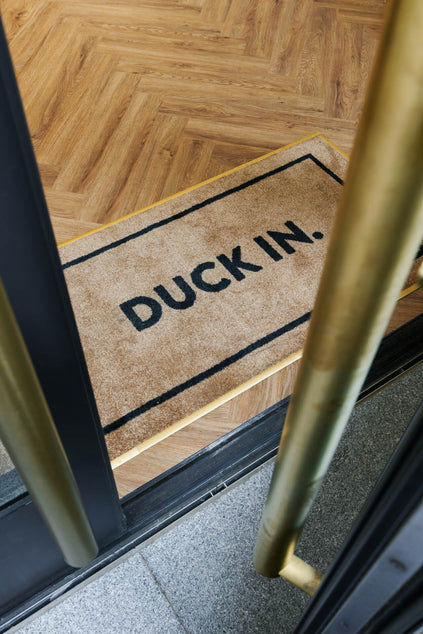 Duck & Dry Mayfair salon doorway with a "Duck In." mat on the herringbone wood floor