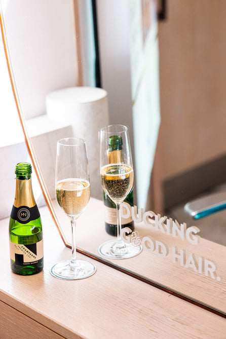 A glass of champagne and a mini bottle at a Duck & Dry salon station in front of a mirror