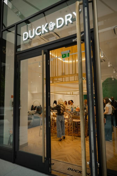 Exterior of a Duck & Dry salon at night, with the lit sign above the glass doors and guests inside